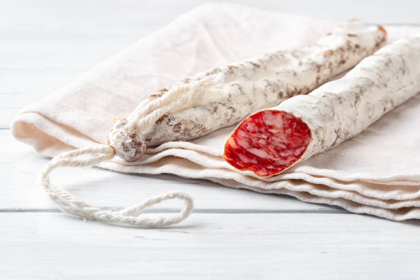 Two thin catalan fuet sausages on a white linen cloth over a wooden table. Close-up of traditional spanish dry-cured pork salami with white noble mold. Tasty meat delicacy. High key image. stock photo