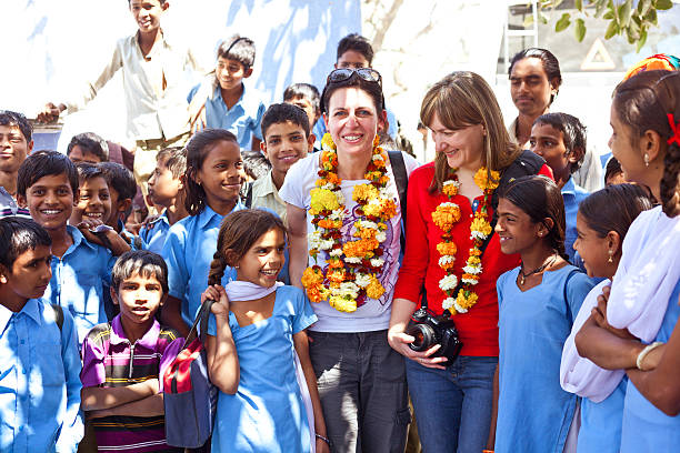 Sabbalpura, India - March 15, 2014: Two female tourists wearing flowers garlands on their necks standing among group of happy indian school girls and boy in the rural village, Rajasthan, India.
