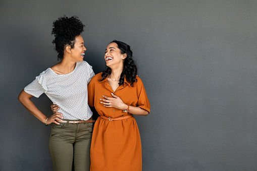 Happy smiling multiethnic women embracing each other against grey wall with copy space. Happy laughing girls standing on gray background and looking at each other. Carefree african american girl having fun with her latin best friend on grey wall with copy space.