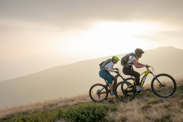 Two women ride up grassy hillside on electric mountain bikes Sun sets over distant mountain range electric bicycle stock pictures, royalty-free photos & images