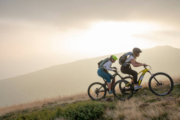 Two women ride up grassy hillside on electric mountain bikes Sun sets over distant mountain range electric bike stock pictures, royalty-free photos & images