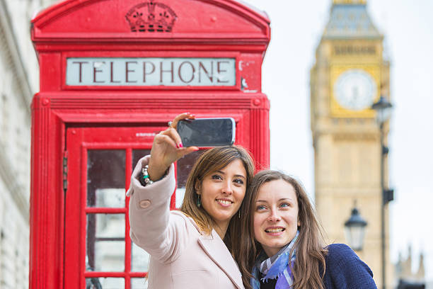 Two women taking a selfie in London. Two beautiful women taking a selfie in London with Big Ben and red phone booth on background. They are in their twenties, holding the phone and looking at it. Focus on the face. red telephone box stock pictures, royalty-free photos & images