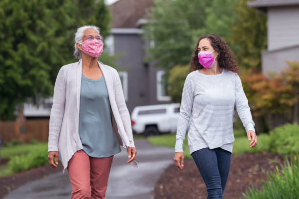 Two women wearing protective face masks enjoying the outdoors during Coronavirus A mixed race senior adult woman is out for a walk with her adult daughter. The two women are wearing protective face masks. They are enjoying the outdoors while taking proper social distancing measures to avoid viral infection of COVID-19. 2 people walking stock pictures, royalty-free photos & images