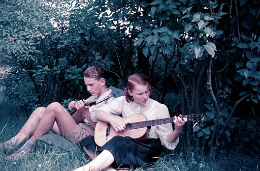 Lower Saxony, Germany, 1957. Two young adults practicing on stringed instruments outside in a park.