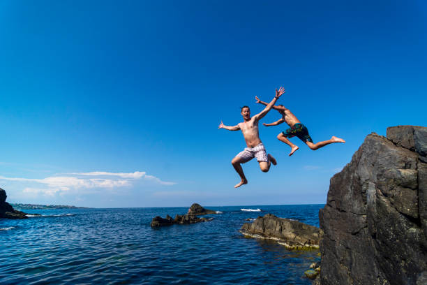 Two young men jumping off cliff into sea Two brave divers jumping off cliff into sea cliff jumping stock pictures, royalty-free photos & images