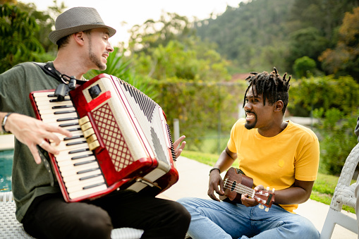 Two diverse young musicians smiling while jamming together outside with an accordion and a ukulele