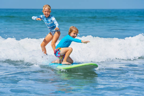 Two young surfers ride with fun on one surfboard Happy baby boy and girl - young surfers ride with fun on one surfboard. Active family lifestyle, kids outdoor water sport lessons, swimming activity in surf camp. Sea beach summer holiday with child. children only photos stock pictures, royalty-free photos & images