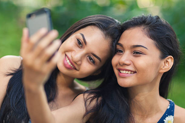 Two young women taking a selfie Two young women outdoors in a park taking a selfie with a smartphone. philippines girl stock pictures, royalty-free photos & images