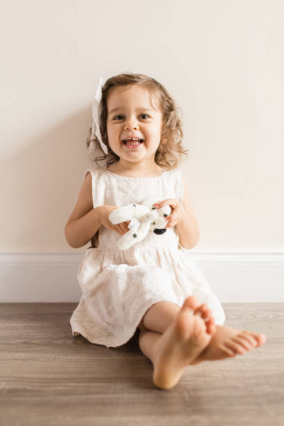 Two-Year-Old Toddler Girl Laughing While Sitting on the Floor in a Cream-Colored Linen Dress & a Bow at Home During COVID-19 Two-Year-Old Toddler Girl Sitting on the Floor in a Cream-Colored Linen Dress & a Bow at Home During COVID-19 beautiful young brunette girl playing with her dog stock pictures, royalty-free photos & images