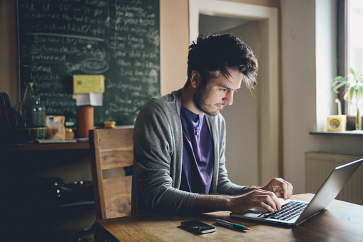 Vintage toned image of a young man working at home, freelancer, designer or writer, checking e-mails or typing on the laptop. Only natural light used, higher iso settings. Interior looks like a modern office, home or a co-working space. 