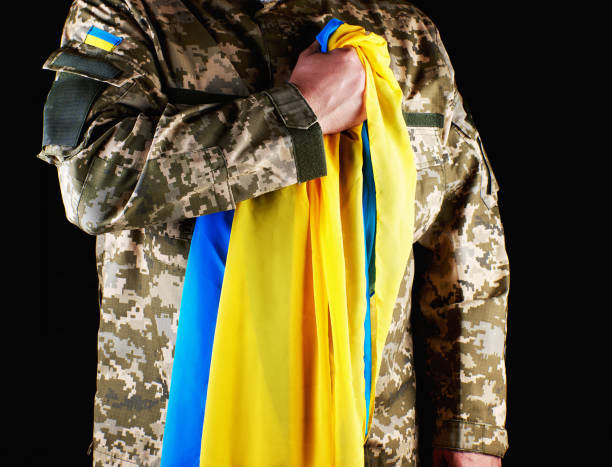soldat ukrainien tient dans sa main le drapeau jaune-bleu de l'état, il a appuyé sa main sur sa poitrine - camouflage ukraine photos et images de collection
