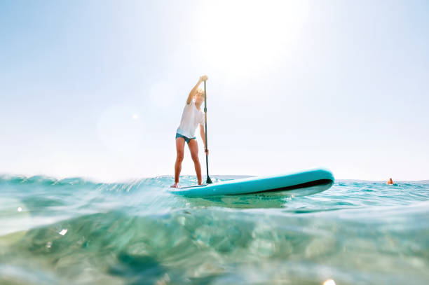 Under the water view angle to the smiling blonde teenager boy rowing stand up paddle board. Active family summer vacation time near the sea concept image. stock photo