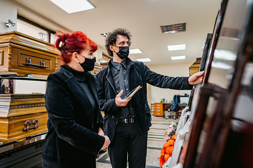 Undertaker showing coffins to a mature widow in funeral parlor. Both of them wearing protective face masks.