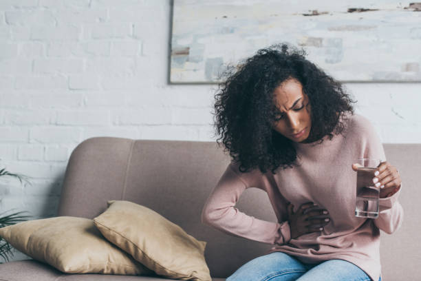 unhappy african american woman holding glass of water while sitting on sofa and suffering from stomach pain unhappy african american woman holding glass of water while sitting on sofa and suffering from stomach pain stomachache stock pictures, royalty-free photos & images