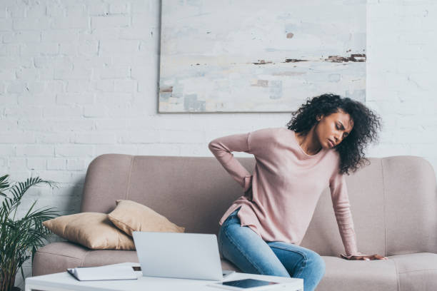 unhappy african american woman suffering from back pain while sitting on sofa near table with digital devices unhappy african american woman suffering from back pain while sitting on sofa near table with digital devices back pain stock pictures, royalty-free photos & images