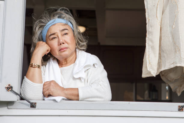 unhappy asian senior woman looking out of window alone at home stock photo