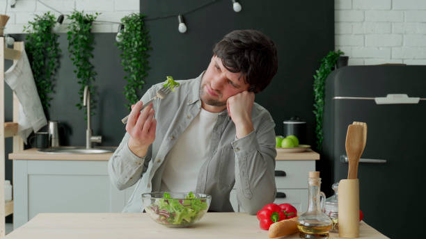 Unhappy man eating vegetable salad at table in kitchen. Displeased young man eating green leaf lettuce Unhappy man eating vegetable salad at table in kitchen. Displeased young man eating green leaf lettuce. disgust photos stock pictures, royalty-free photos & images
