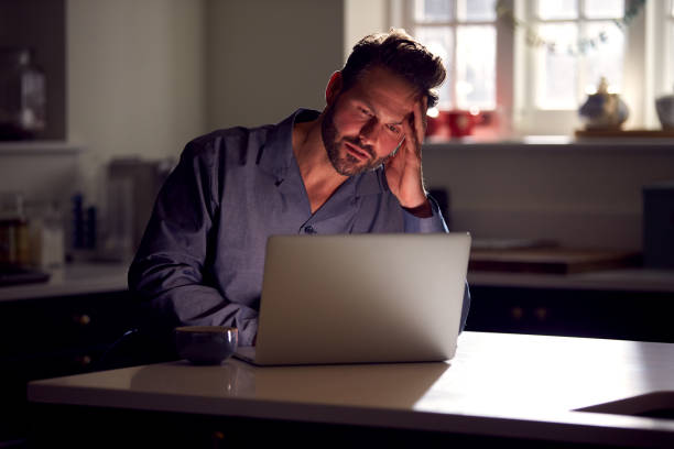 Unhappy Man Wearing Pyjamas Sitting In Kitchen At Night Using Laptop Unhappy Man Wearing Pyjamas Sitting In Kitchen At Night Using Laptop fomo stock pictures, royalty-free photos & images