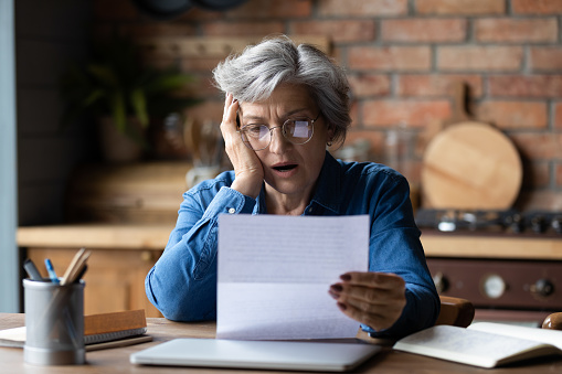 Unhappy mature wearing glasses reading bad news in letter, stressed grey haired female with open mouth looking at paper sheet, sitting at desk, shocked by negative message in correspondence