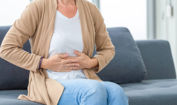 Unhappy woman stomach ache, mature woman with stomach pain feeling unwell sitting in living room stock photo