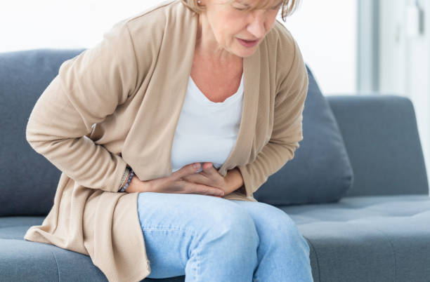 Unhappy woman stomach ache, mature woman with stomach pain feeling unwell sitting in living room stock photo