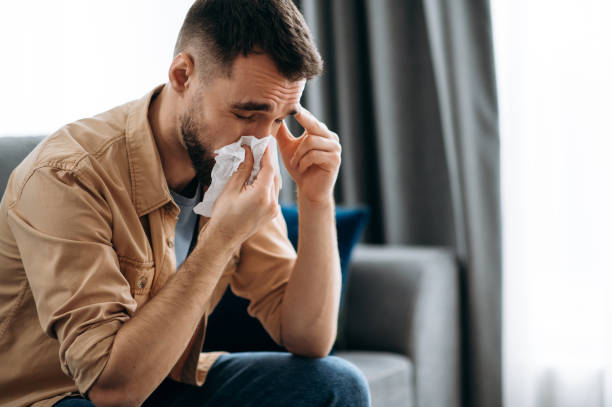 Unhealthy young man. Caucasian guy is sick with flu, he sneezes and blows his nose in a napkin while sitting on the couch at home stock photo