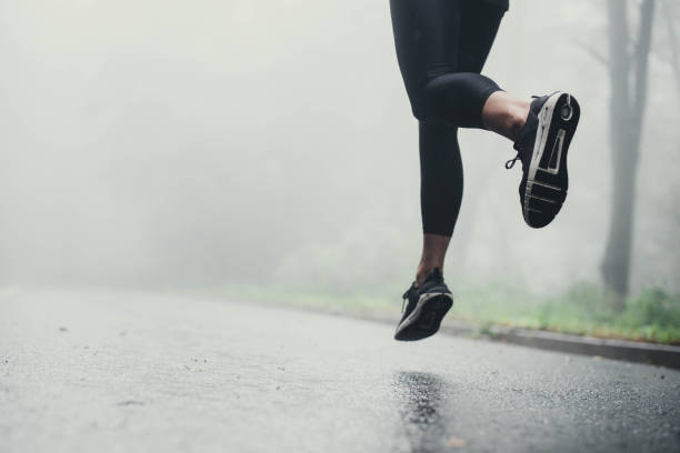 Unrecognizable athlete jogging on the road during rainy day. Unrecognizable athletic woman running on the road during foggy day in nature. Copy space. running stock pictures, royalty-free photos & images
