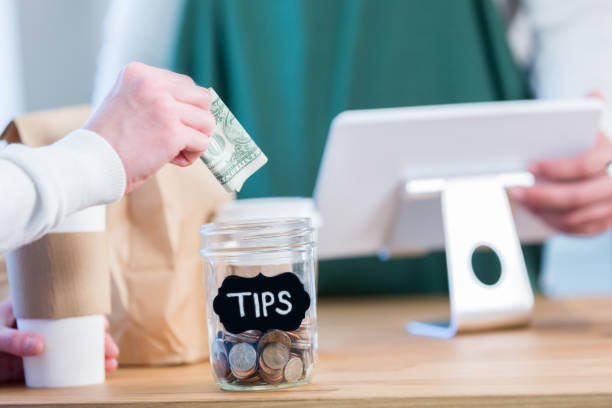 Unrecognizable coffee shop customer using tip jar An unrecognizable coffee shop customer stands across the checkout counter from an unrecognizable barista and reaches out to put a paper bill in the tips jar. tipping in restaurants stock pictures, royalty-free photos & images