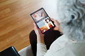 Unrecognizable elderly woman with gray hair is sitting on sofa in her living room and reading about Covid-19 new treatments and news on the digital tablet