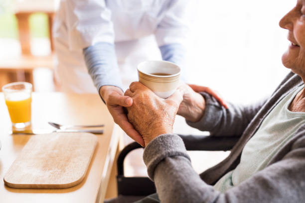 Unrecognizable health visitor and a senior woman during home visit. Unrecognizable health visitor and a senior woman during home visit. A nurse giving tea to an elderly woman sitting at the table. Close up. home caregiver stock pictures, royalty-free photos & images