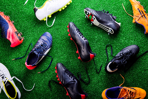 Unrecognizable little football player with soccer ball tying shoelaces, against artificial grass. Studio shot on green grass.
