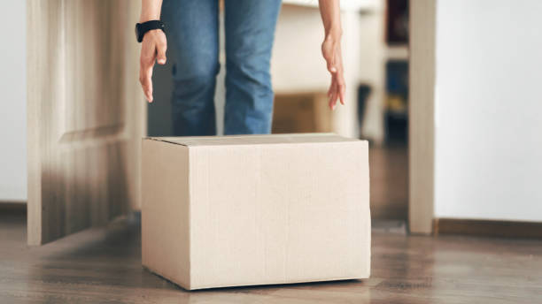 Unrecognizable person picking cardboard box up from the floor Quarantine Delivery. Closeup of unrecognizable person taking and picking up carton package from the floor standing at home near door. Selective focus on cardboard box and hands, blurred background image technique stock pictures, royalty-free photos & images