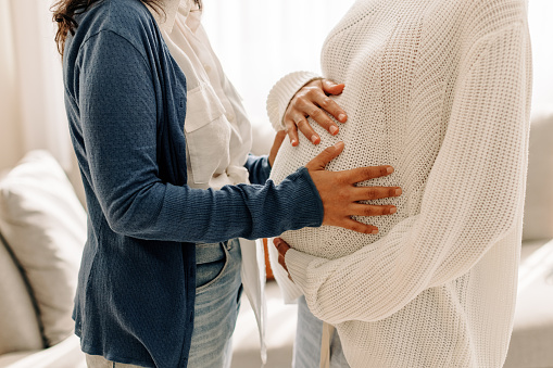 Unrecognizable young woman touching a surrogate mother's belly bump. Young woman spending time with her surrogate at home. Woman feeling the movement of a pregnant woman's baby.