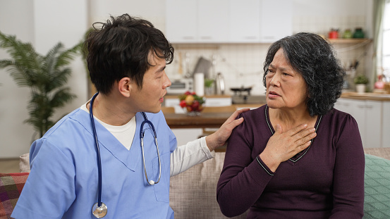unwell asian senior female patient describing her issue to male nurse during home visit in the living room. she touches her chest as the man asking about details