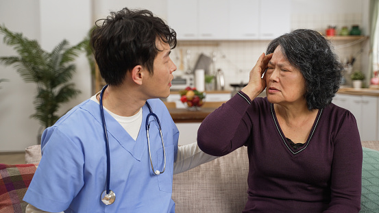 unwell asian senior female patient describing her issue to male nurse during home visit in the living room. she touches her head as the man asking about details