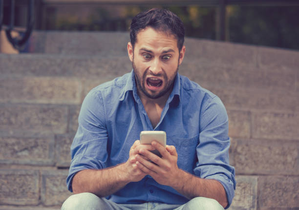 Upset man looking at his mobile phone sitting on steps outdoors Shocked upset young man looking at his mobile phone seeing bad news or reading text message sitting on stairs outside corporate building. Human emotion, reaction, expression worried man funny stock pictures, royalty-free photos & images