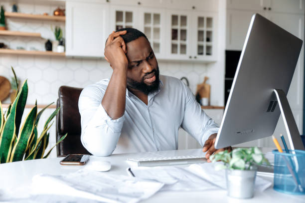Upset sad young african american man, manager, broker or freelancer, working at home at the computer, experiencing stress at work, holding his head with his hand, looking sadly at the screen stock photo