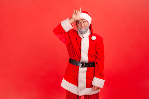 Upset unlucky elderly man with gray beard wearing santa claus costume showing looser gesture holding finger near forehead, depressed with her fail. Indoor studio shot isolated on red background.