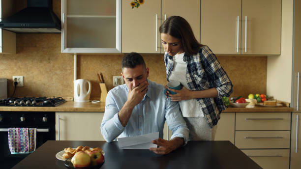 Upset young man reading unpaid bills and hugged by his wife supporting him in the kitchen at home stock photo