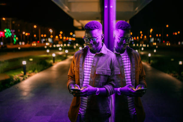 Using Phone in a front of neon lights on the street Young handsome black man using phone, typing text message in a front of neon blue light office window in rainy night only men photos stock pictures, royalty-free photos & images
