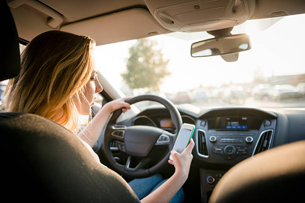 Using phone while driving Young woman looking to her smartphone while driving car - rear view, sun shines through front window distracted stock pictures, royalty-free photos & images