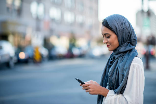 Using Technology A Muslim woman is outdoors on a sunny day. She is wearing casual clothing and a head scarf. She is standing near a road and sending a message with her smartphone. west asian ethnicity stock pictures, royalty-free photos & images