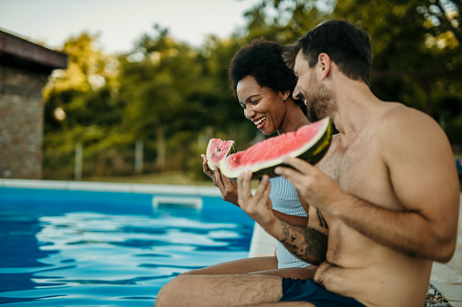 Spontaneous photo of a young adult, Mixed raced couple, enjoying in swimming pool, laughing and eating watermelon. Both radiating love and enjoyment on a beautiful sunny vacation time