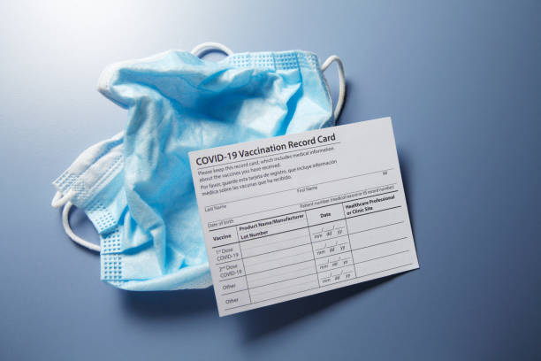 COVID-19 Vaccination Card On Discarded Protective Face Mask A blank COVID-19 vaccination record card rests on top of a discarded protective face mask on a blue background. cdc vaccine card stock pictures, royalty-free photos & images