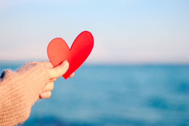 Valentine day concept, soft focus woman hand holding red heart, blurred sea background. Card with a carved red paper heart in a man's hand against a background of beautiful azure blue sea ocean. stock photo