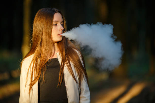 Vape teenager. Young cute girl in casual clothes smokes an electronic cigarette outdoors in the forest at sunset in summer. Bad habit. Vape teenager. Young cute girl in casual clothes smokes an electronic cigarette outdoors in the forest at sunset in summer. Bad habit that is harmful to health. Vaping activity. little girl smoking cigarette stock pictures, royalty-free photos & images