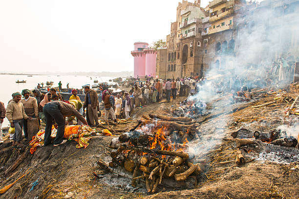 Varanasi burning grounds Varanasi, India - January 5, 2014: Burning Ghats and the cremation grounds on the bank of river Ganges in Varanasi. People, man, standing around deceased, covered with orange sheet and decorated with colorful ribbons.In front view burning corpses.  ghat stock pictures, royalty-free photos & images