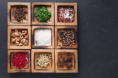 Rustic wooden box with many compartments filled with seasonings. Salt, peppers, coriander seeds, cloves, walnuts and green parsley on black background. Flavorings and condiments for food and drinks
