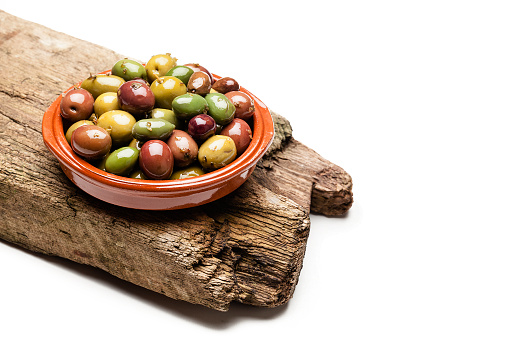 Brown clay bowl filled with various types of fresh olives placed on a rustic wood strip isolated on white background. Predominant colors are green and brown. Useful copy space available for text and/or logo. High key DSRL studio photo taken with Canon EOS 5D Mk II and Canon EF 100mm f/2.8L Macro IS USM.