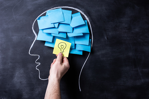 Head shape drown on a blackboard with many blue post-it notes forming the brain. There is a hand showing one yellow note with a light bulb drawn on it, meaning solution or bright idea.